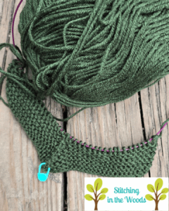 Forest green yarn and a part of a knitted afghan square is laying on a wooden deck, you can see the circular needle cable running through the active row